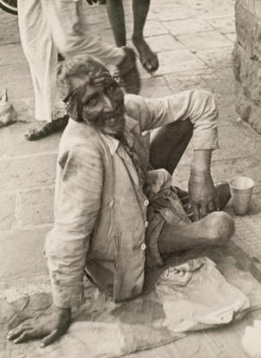 (Emaciated man with headscarf on ground with cup)