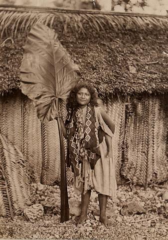 Girl with baby on her back, with large taro leaf outside hut